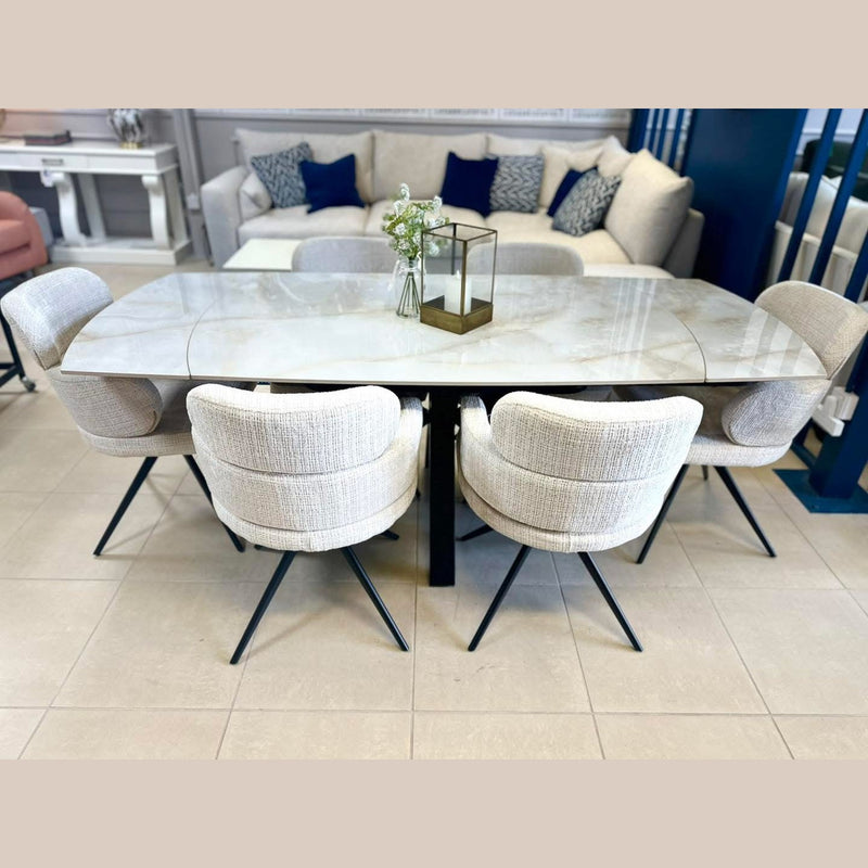A modern dining room features a marble-top rectangular table with black metal legs, six Orbit Dining Chairs in beige fabric, a glass vase with flowers, and a brass lantern centerpiece. In the background is a sofa with blue and white pillows.