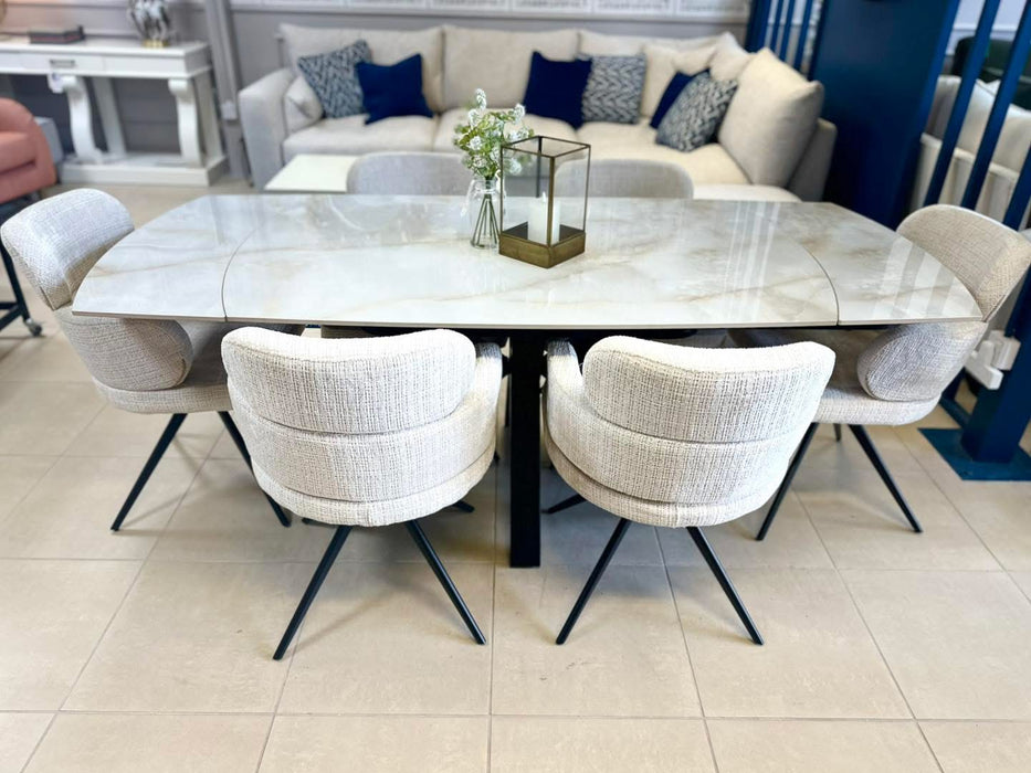 A modern dining room features a marble-top rectangular table with black metal legs, six Orbit Dining Chairs in beige fabric, a glass vase with flowers, and a brass lantern centerpiece. In the background is a sofa with blue and white pillows.