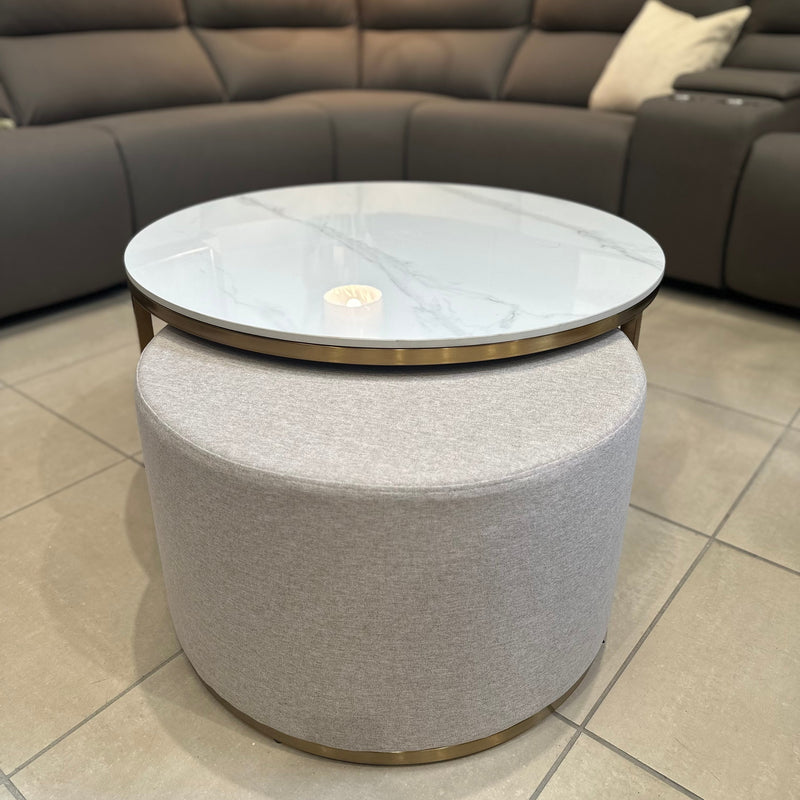 The Vienna Coffee Table + Footstool features a marble top with gold accents and a light gray upholstered ottoman beneath, placed in front of a modern dark gray sectional on tiled floors.