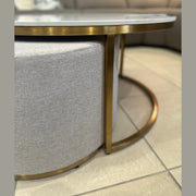 The Vienna Coffee Table + Footstool, featuring a gold metal frame and light gray upholstered ottoman, stands on a tiled floor with a cushioned sofa in the background.