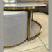 The Vienna Coffee Table + Footstool, featuring a gold metal frame and light gray upholstered ottoman, stands on a tiled floor with a cushioned sofa in the background.