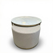 The Vienna Coffee Table + Footstool boasts a round marble top with glass overlay, a beige upholstered base, and gold accents. Its clean lines and minimalist style make it an elegant focal point against any white backdrop.