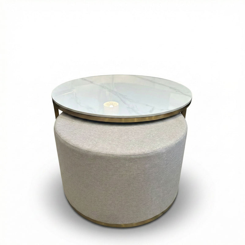 The Vienna Coffee Table + Footstool boasts a round marble top with glass overlay, a beige upholstered base, and gold accents. Its clean lines and minimalist style make it an elegant focal point against any white backdrop.