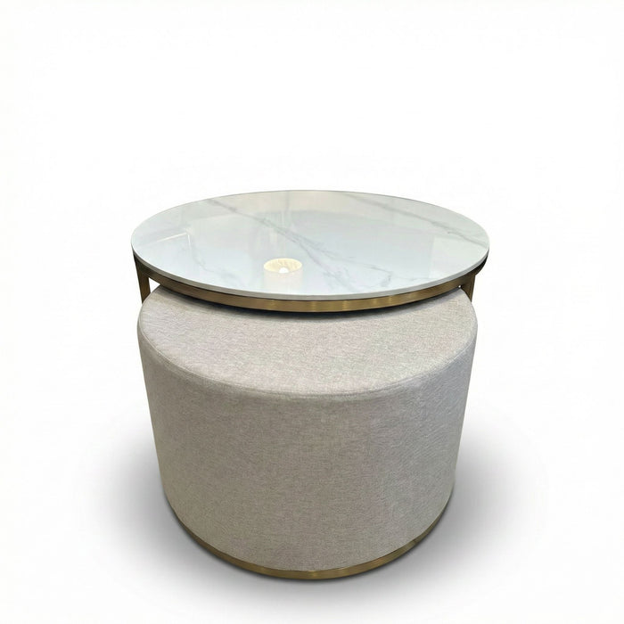 The Vienna Coffee Table + Footstool boasts a round marble top with glass overlay, a beige upholstered base, and gold accents. Its clean lines and minimalist style make it an elegant focal point against any white backdrop.