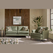 A modern living room featuring the Joni Range sofa and matching armchair, patterned cushions, a potted plant, side table, pouf, and wooden accent wall with abstract art. Sunlight streams in—perfect for contemporary homes.