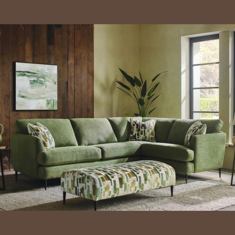 A modern living room featuring the Joni Range: a compact green corner sofa, matching ottoman, patterned pillows, wooden wall panels, large windows, potted plant, table lamp, and abstract green artwork—ideal for contemporary homes.