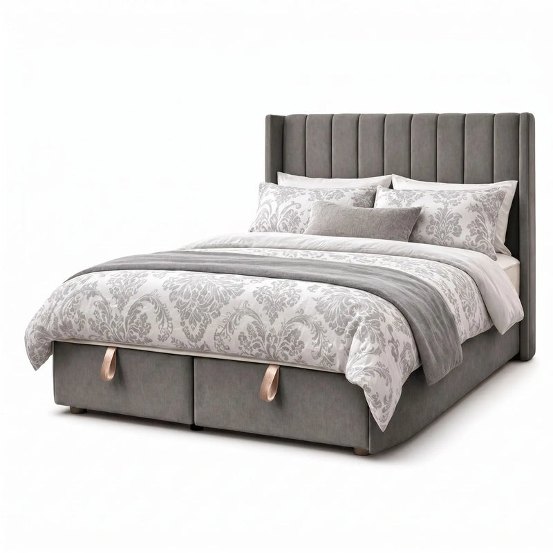 The Hilton Storage Bedframe features a grey upholstered finish, a tall vertical-channeled winged headboard, two storage drawers with beige handles at the base, plus white and grey patterned bedding with several pillows.