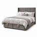 The Hilton Storage Bedframe features a grey upholstered finish, a tall vertical-channeled winged headboard, two storage drawers with beige handles at the base, plus white and grey patterned bedding with several pillows.