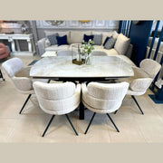 A modern dining area showcases the Positano Table + 6 Orbit Chairs set, topped with a glass vase of white flowers and a candleholder, while sofas and decor can be seen in the background.