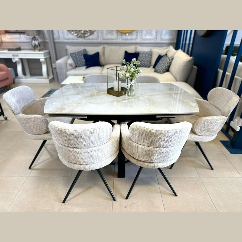 A modern dining area showcases the Positano Table + 6 Orbit Chairs set, topped with a glass vase of white flowers and a candleholder, while sofas and decor can be seen in the background.