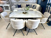 A modern dining area showcases the Positano Table + 6 Orbit Chairs set, topped with a glass vase of white flowers and a candleholder, while sofas and decor can be seen in the background.