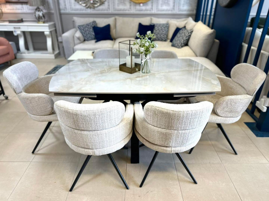 A modern dining area showcases the Positano Table + 6 Orbit Chairs set, topped with a glass vase of white flowers and a candleholder, while sofas and decor can be seen in the background.