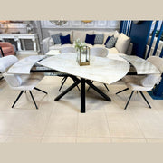 The Orbit Dining Chair, with light fabric and black metal legs, surrounds a glass-top table featuring a geometric white marble base and black legs in a modern showroom display.