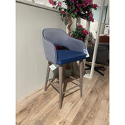 The Turim Counter Stool, featuring light wood legs, a blue cushioned seat, and a textured gray backrest, is set on a wooden floor near a partial wall and a potted plant with pink flowers.