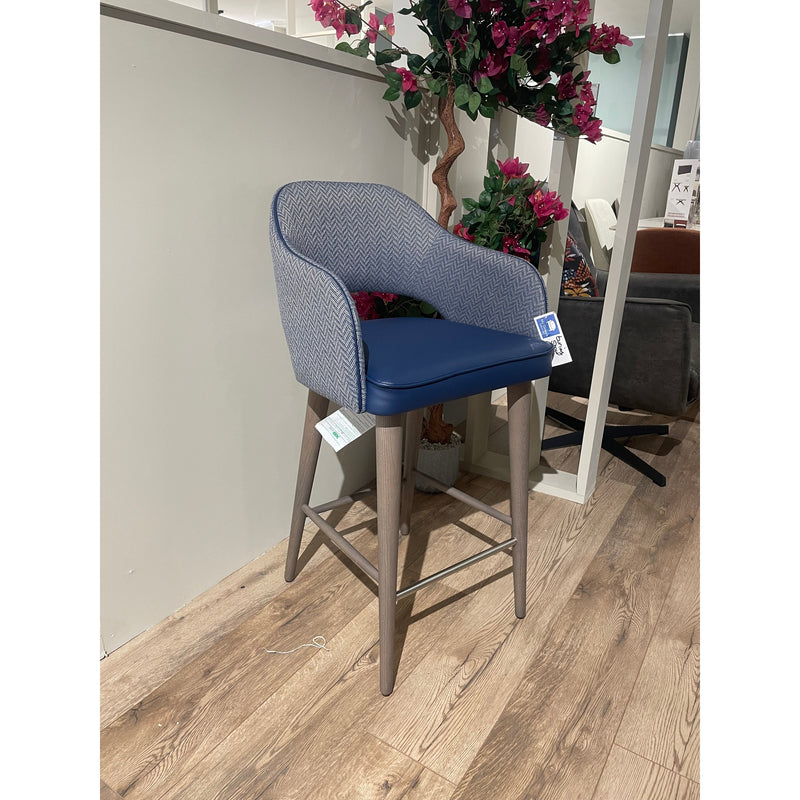 The Turim Counter Stool, featuring light wood legs, a blue cushioned seat, and a textured gray backrest, is set on a wooden floor near a partial wall and a potted plant with pink flowers.