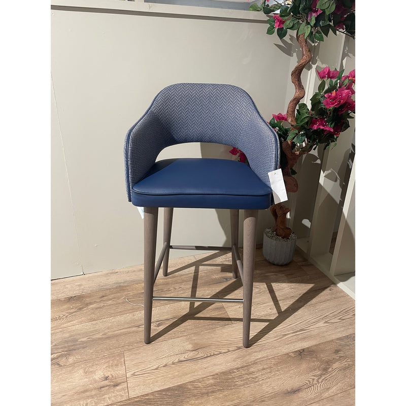 The Turim Counter Stool features a blue cushioned seat, curved fabric backrest, and light wooden legs. Shown on a light wood floor beside a potted plant with pink flowers.