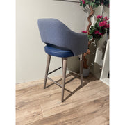 The Turim Counter Stool, featuring a blue cushioned seat and backrest with wooden legs and a footrest, stands on a wooden floor beside a potted artificial plant with pink flowers against a beige wall.