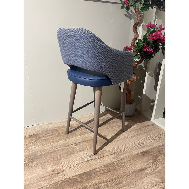The Turim Counter Stool, featuring a blue cushioned seat and backrest with wooden legs and a footrest, stands on a wooden floor beside a potted artificial plant with pink flowers against a beige wall.