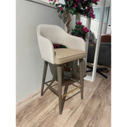 The Turim Counter Stool features a beige cushioned seat, a light chevron-patterned backrest, and wooden legs with a brass footrest, shown on a wood floor near a white wall and decorative plant.