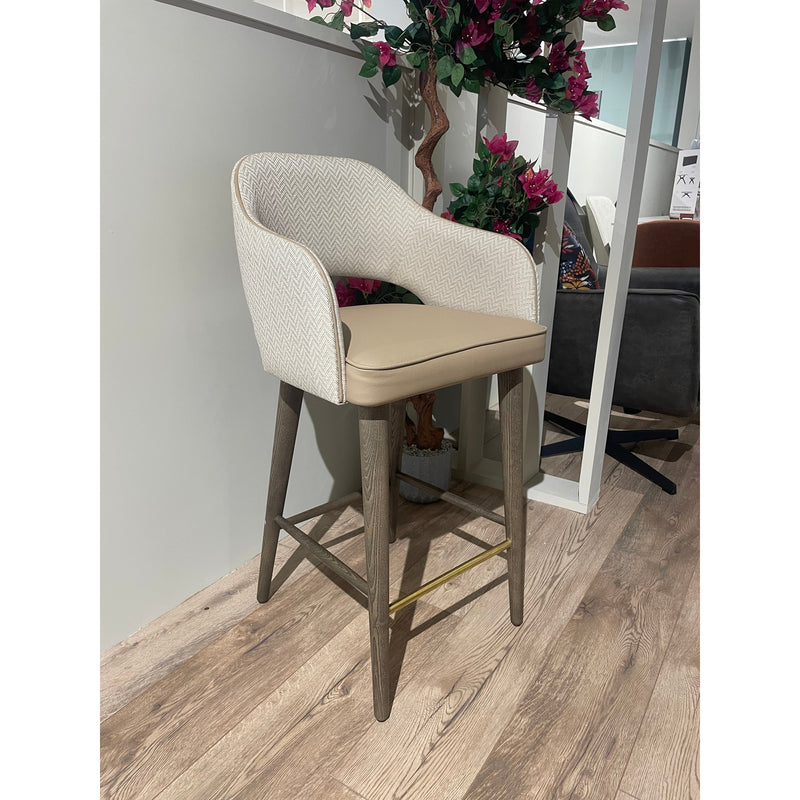 The Turim Counter Stool features a beige cushioned seat, a light chevron-patterned backrest, and wooden legs with a brass footrest, shown on a wood floor near a white wall and decorative plant.