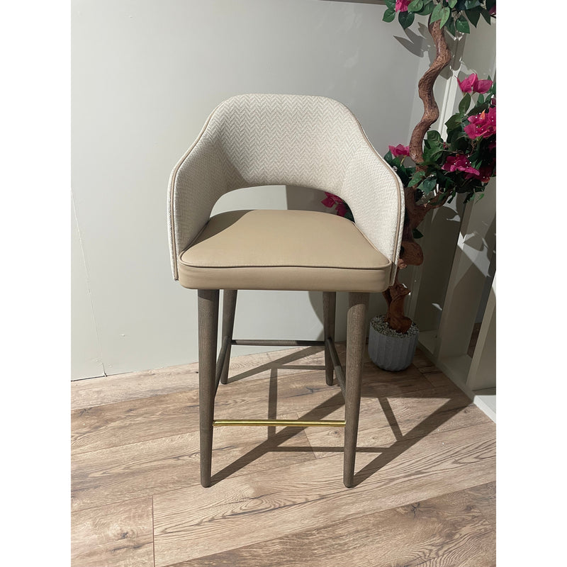 The Turim Counter Stool, featuring a modern beige cushioned seat and backrest, wooden legs, and a gold footrest, stands on a light wood floor beside a potted plant with pink flowers.