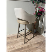 The Turim Counter Stool, featuring a curved backrest and wooden legs, sits on a light wood floor beside a small artificial pink-flowered tree against a light-colored wall.