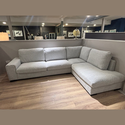 The Sorrento Chaise Corner, with modern stone-coloured textured upholstery, sits on a wooden floor in a showroom, framed by art on dark walls in the background.