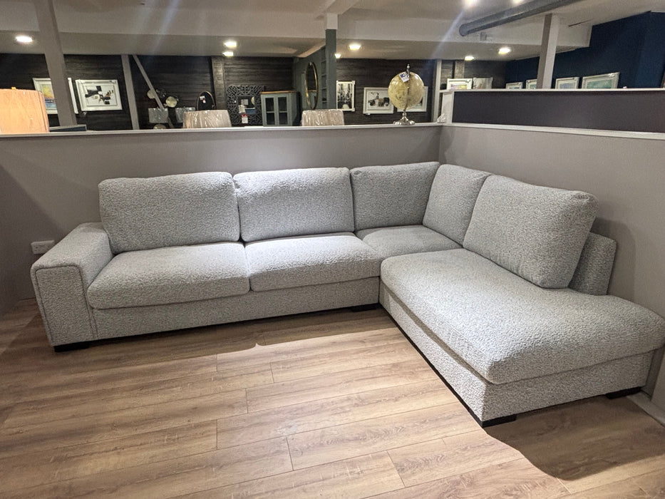 The Sorrento Chaise Corner, with modern stone-coloured textured upholstery, sits on a wooden floor in a showroom, framed by art on dark walls in the background.