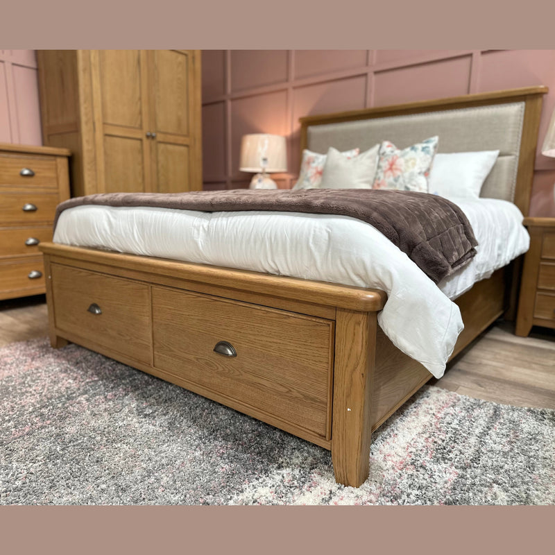 The Aylesbury bedframe, featuring under-bed storage drawers, is styled with white bedding, a brown blanket, and floral pillows in a cozy bedroom with matching wooden furniture and soft pink paneled walls.