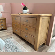 The Aylesbury wide chest, featuring six drawers with silver cup handles in solid smoked oak, stands against a pink wall next to a matching three-drawer bedside table topped with a lamp and plant. Part of the bed and a rug are also visible.