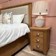 A wooden bed with a beige upholstered headboard, white bedding, and floral pillows sits next to the Aylesbury locker for elegant storage, topped with a white table lamp wrapped in plastic against a pink paneled wall and wooden floor.