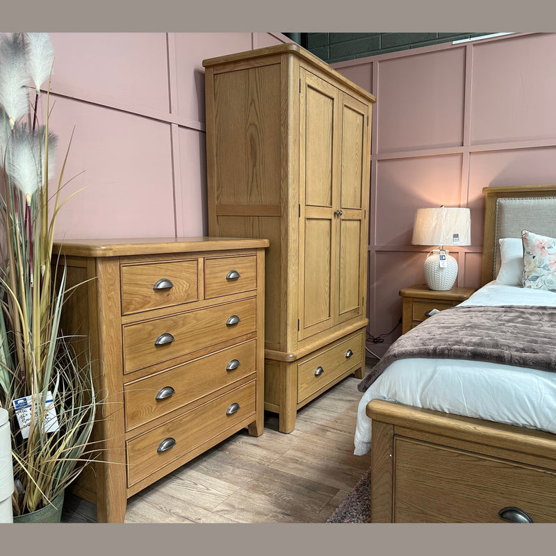 A cozy bedroom with elegant furniture, featuring the Aylesbury tall chest in smoked oak with silver cup handles, a matching wardrobe, and a bed with a beige headboard. Soft white bedding, pastel floral pillows, and a brown throw complete the inviting look.