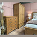 A cozy bedroom with elegant furniture, featuring the Aylesbury tall chest in smoked oak with silver cup handles, a matching wardrobe, and a bed with a beige headboard. Soft white bedding, pastel floral pillows, and a brown throw complete the inviting look.