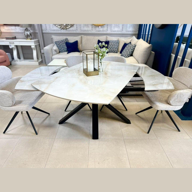 A modern dining room with a Positano Swivel Dining Table featuring a triangular ceramic top and black metal legs, surrounded by four beige upholstered chairs. A gold lantern and a flower vase sit on the table, with a gray sofa in the background.