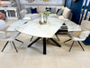 A modern dining area with a Positano Table and 6 Orbit Chairs, featuring a triangular sintered stone top, black legs, and light gray upholstery. A glass and gold vase with flowers is centered, with a beige sofa in the background.