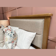 Aylesbury bedframe in light oak with an upholstered headboard and nailhead trim, topped with white pillows and a floral cushion. A lamp sits on the bedside table against a pink paneled wall.