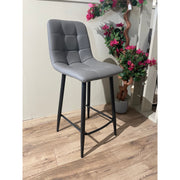 A black steel bar stool with a leatherette seat in gray, standing in a room with a potted plant and wooden flooring.