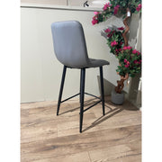 The Fredrick bar stool, featuring a gray leatherette seat and black steel legs, sits on a wood-patterned floor near a white wall and a potted artificial plant with green leaves and pink flowers.
