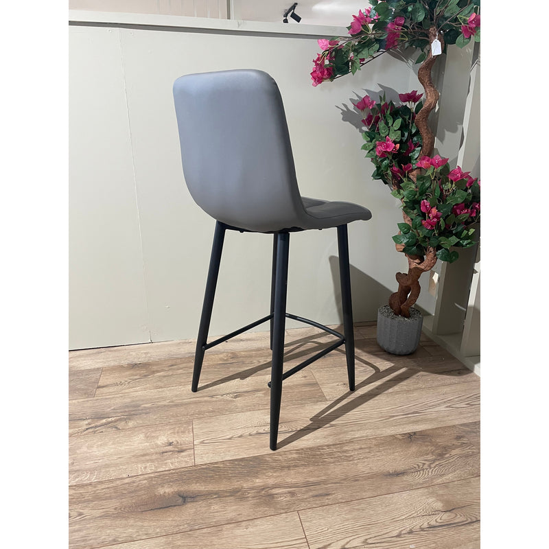 The Fredrick bar stool, featuring a gray leatherette seat and black steel legs, sits on a wood-patterned floor near a white wall and a potted artificial plant with green leaves and pink flowers.