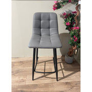 The Fredrick bar stool, featuring a gray leatherette seat, square-tufted backrest, and black steel legs, sits on a wooden floor beside a potted plant with pink flowers against a light-colored wall.