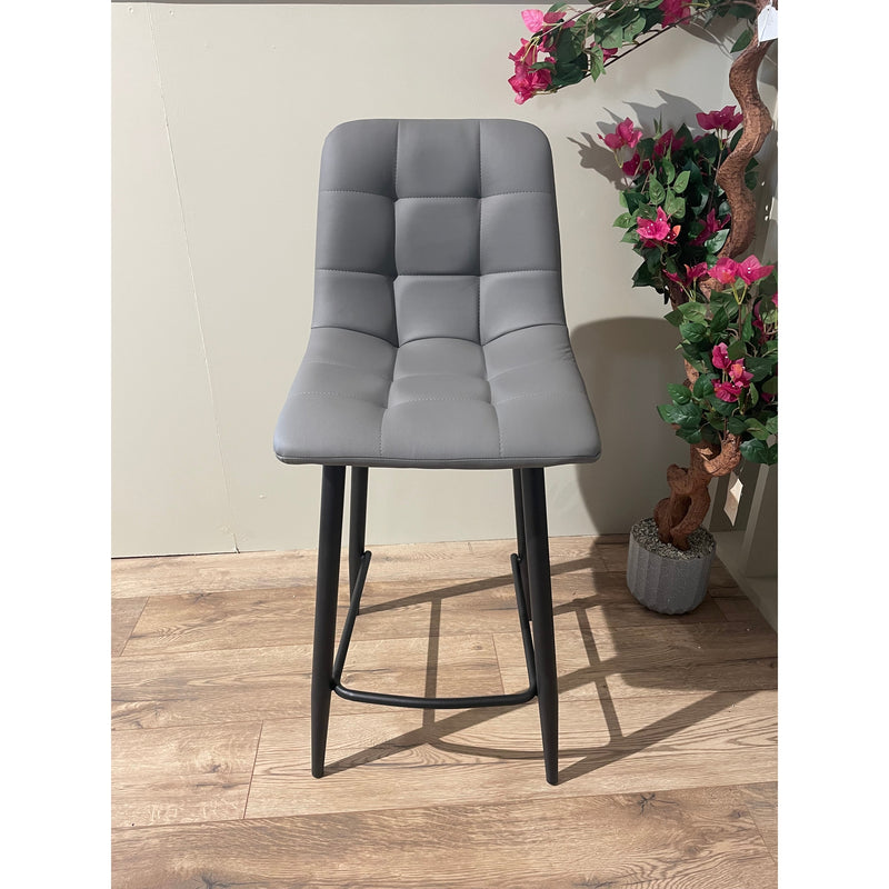 The Fredrick bar stool, featuring a gray leatherette seat, square-tufted backrest, and black steel legs, sits on a wooden floor beside a potted plant with pink flowers against a light-colored wall.