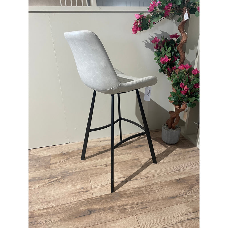The George bar stool, featuring a light grey cushioned seat and black metal legs, stands on a wooden floor beside a potted plant with pink flowers. A white price tag hangs from the stool.