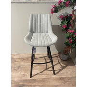 The George bar stool, featuring a contemporary light gray upholstered seat with vertical stitching and black metal legs, stands on a wooden floor beside a potted artificial plant with pink flowers.