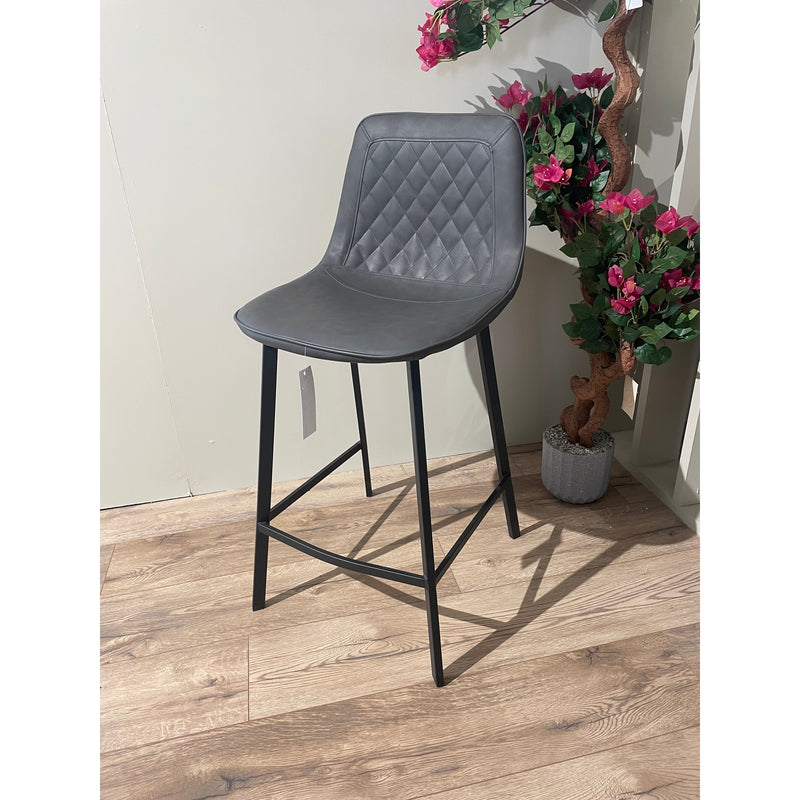 A gray upholstered retro bar stool with a diamond pattern on the backrest, positioned on a wooden floor against a wall, with a potted plant in the background.