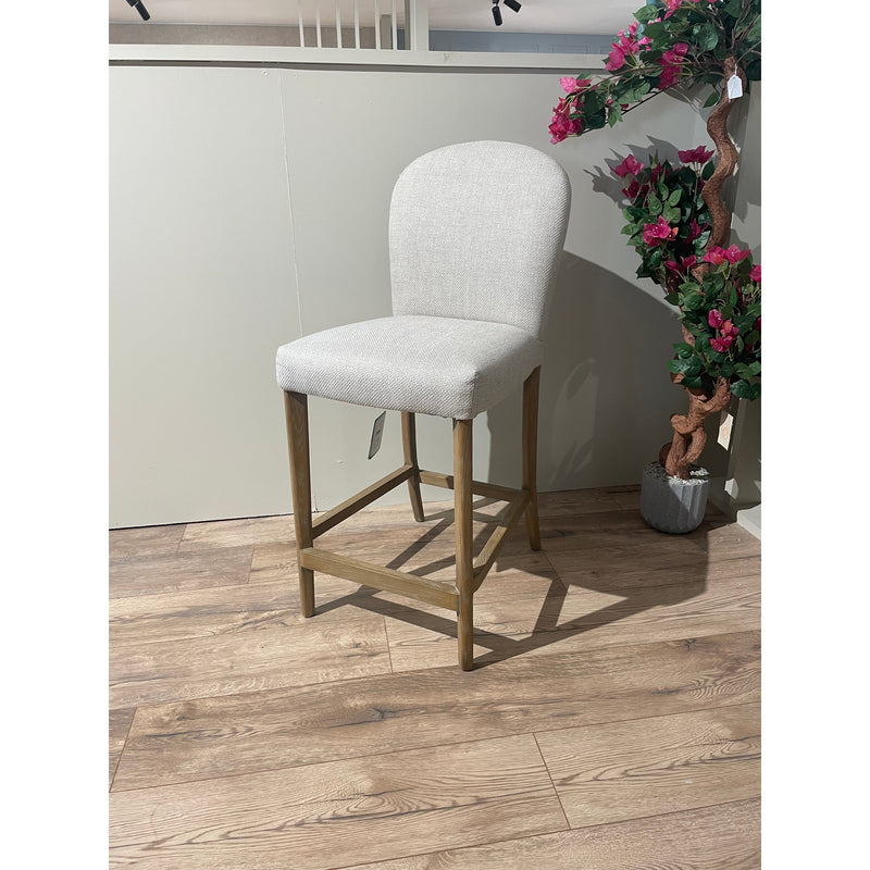 The Jenny Counter Stool, upholstered in cream fabric with a wooden frame, sits on a wood floor beside a potted artificial plant with pink flowers. A pale wall and railing are visible in the background.