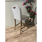 The Jenny Counter Stool, featuring light gray upholstery, wooden legs, nailhead trim, and a brown leather handle, sits on a wooden floor beside a potted artificial plant with pink flowers and a cream partition wall.