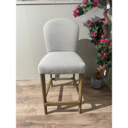 The Jenny Counter Stool features light gray upholstery, a curved backrest, wooden legs, and a stylish brown leather handle. It sits on a wood-patterned floor beside a potted plant with pink flowers.