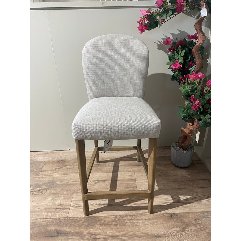 The Jenny Counter Stool features light gray upholstery, a curved backrest, wooden legs, and a stylish brown leather handle. It sits on a wood-patterned floor beside a potted plant with pink flowers.