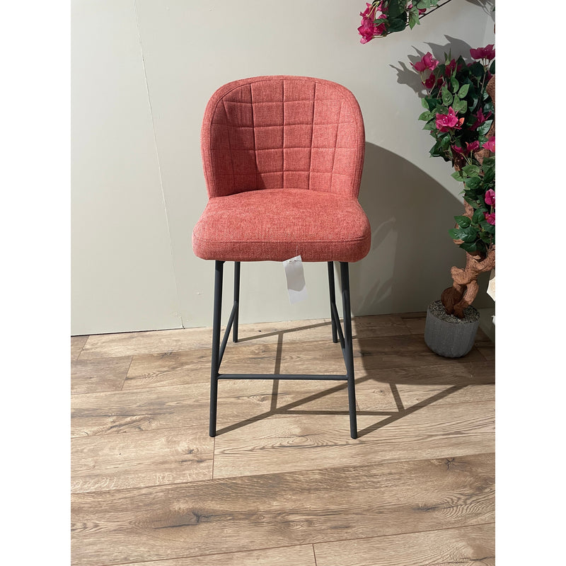 The Clio bar stool showcases a red upholstered seat and black metal legs, complemented by a white tag on the seat. Pink floral plants in gray pots accent the nearby light wall above the wooden floor.