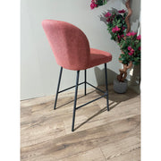 The Clio bar stool features a modern design with a curved red upholstered backrest and black metal legs, placed on a wooden floor beside a potted plant with pink flowers against a light gray wall.